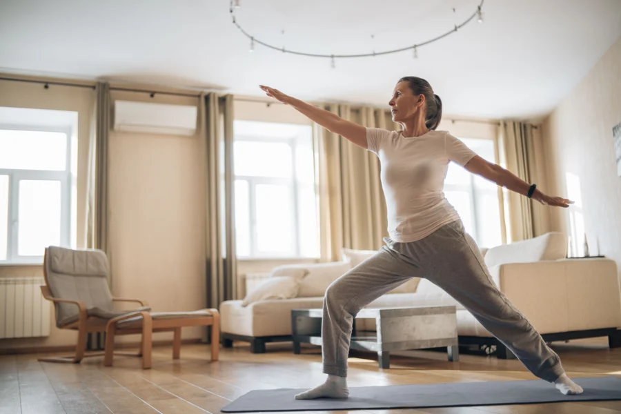 Femme d’âge mûr debout dans un salon, jambes écartées, bras tendus, pendant un exercice à la maison